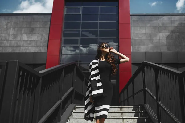 穿着黑色衣服、戴着太阳镜的漂亮、时髦的黑发模特站在现代建筑前 — 图库照片