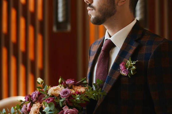 Vágott portré egy felismerhetetlen jóképű férfiról, nyakkendőben és elegáns kockás öltönyben, boutonniere-rel, kezében egy nagy csokor friss virággal. Stílusos vőlegény várja a menyasszonyát. — Stock Fotó