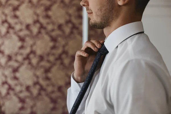 Felismerhetetlen jóképű férfi fehér ingben nyakkendőt vesz fel. Az elegáns vőlegény reggele az esküvő előtt — Stock Fotó