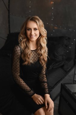 Siyah bir kokteyl elbisesi giymiş, gülümseyen ve içeride poz veren genç model bir kadın.