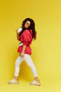 Pembe kapüşonlu, beyaz pantolonlu, elinde kağıt kahve fincanı olan ve sarı arka planda izole edilmiş içecekten hoşlanan şık model kadının tam boy stüdyo fotoğrafı.