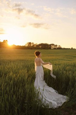 Beyaz elbiseli güzel, mutlu esmer gelin gün batımı çayırında arkası ile poz veriyor.