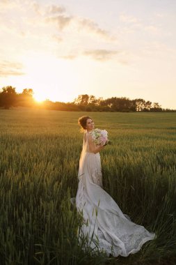 Beyaz dantelli gelinlik içindeki mutlu gelin gün batımında çayırda çiçek demeti ile poz veriyor.