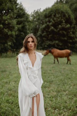 Beyaz cüppeli güzel, şehvetli bir kız yaz çayırında atlarla poz veriyor.