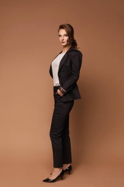 Şık takım elbiseli ve beyaz bluzlu genç bir kadın bej arka planda poz veriyor.