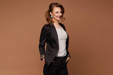 Şık takım elbiseli ve beyaz bluzlu genç bir kadın bej arka planda gülüyor ve poz veriyor.