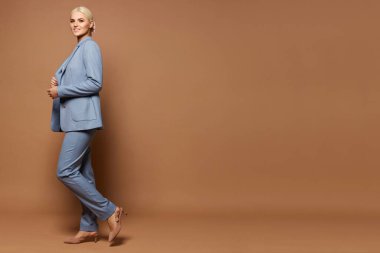 Modayı takip eden mavi takım elbiseli ve bej ayakkabılı genç bir kadın bej arka planda poz veriyor.