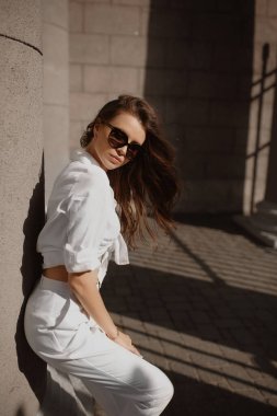 Moda güneş gözlüğü, beyaz pantolon ve gömlek giyen, Avrupa sokaklarında poz veren mükemmel ince vücutlu genç bir kadın model.