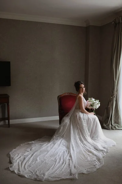 Una mujer joven en un vestido de novia vintage con un ramo de flores se sienta en la silla en el interior vintage — Foto de Stock