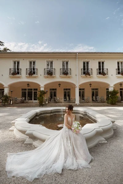 Молода щаслива наречена в розкішній весільній сукні сидить біля фонтану у дворі розкішної вілли в літній день — стокове фото