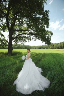 Gelinlik giymiş genç bir kadın. Gelinlik çiçekli. Yaz günü yeşil alanda yalnız başına poz veriyor.