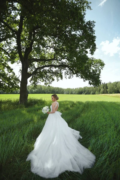 Модель дівчини в розкішній весільній сукні з весільним квітковим букетом, що позує в зеленому полі біля самотнього дерева в літній день — стокове фото