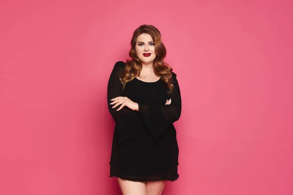 Jovem plus size mulher modelo caucasiano em vestido preto mãos dobradas e posando sobre fundo rosa, isolado com espaço de cópia — Fotografia de Stock