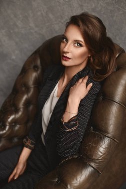 Moda portresi, şık bir takım elbise ve bluzla lüks bir deri koltukta poz veren mükemmel makyaj ve saç stili olan genç bir kadın.