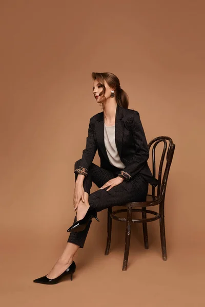 Vuxen kvinna, kaukasiska affärskvinna i grå kostym, blus och svarta skor poserar på stolen över beige bakgrund, isolerad med kopia utrymme — Stockfoto