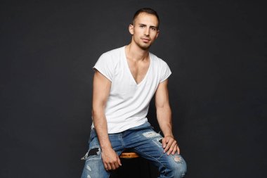 Siyah arka planda beyaz tişörtlü ve kot pantolonlu neşeli bir genç. Karanlık arka planda poz veren günlük kıyafetli yakışıklı bir yetişkin..