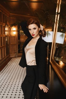 Mükemmel makyajlı, kırmızı dudaklı, saten bluz giyen ve lüks bir iç mekanda poz veren genç bir modelin portresi.