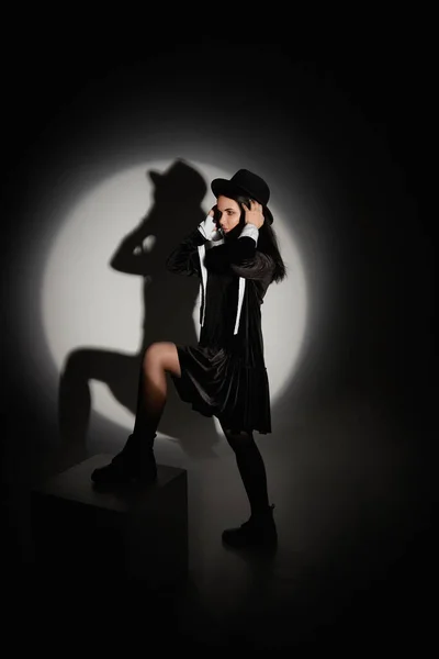 Junge Model-Frau mit schwarzem Hut und kurzem schwarzen Kleid posiert im Scheinwerferlicht im schwarzen Raum — Stockfoto
