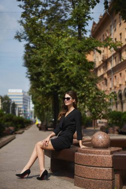 Siyah elbiseli, güneş gözlüklü, uzun bacaklı bir model kız bankta oturuyor ve güneşli bir yaz gününün tadını çıkarıyor.