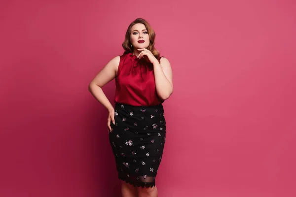 Jovem plus size mulher modelo caucasiano em blusa de cetim vermelho e saia posando sobre fundo rosa. Linda mulher gorda em traje modish sobre fundo rosa — Fotografia de Stock