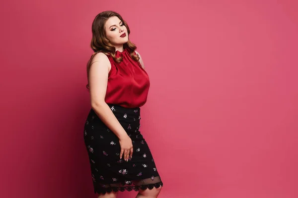 Jovem modelo plus-size menina em blusa de cetim vermelho e saia posando sobre fundo rosa. Pensive mulher gordinha em roupa modish sobre fundo rosa — Fotografia de Stock