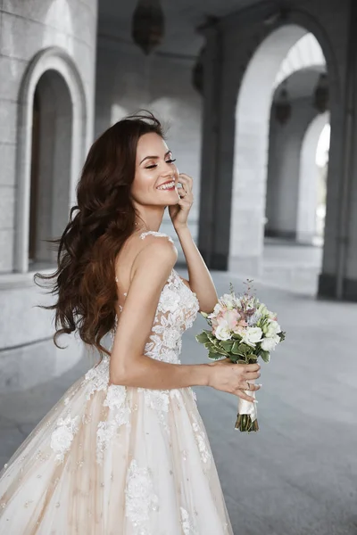 Uma jovem noiva feliz com um penteado de casamento em renda branca no pátio de uma igreja antiga. Uma menina modelo bonita segura um buquê de noiva e posa na cidade velha — Fotografia de Stock