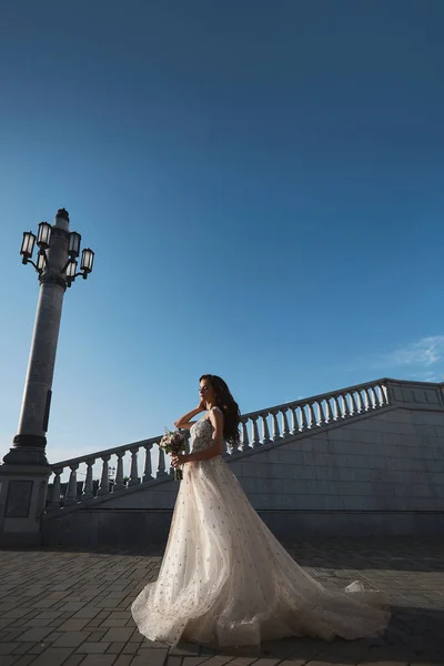 Eine wunderschöne junge Braut in einem luxuriösen Brautkleid aus Spitze posiert im Freien. Romantisches Porträt einer attraktiven brünetten Frau mit hellem Make-up und Hochzeitsfrisur — Stockfoto