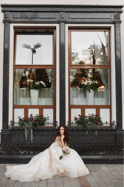 Uma bela noiva jovem vestindo um luxuoso vestido de noiva rendas posando com um buquê de noiva ao ar livre. Retrato romântico ao ar livre de uma menina morena atraente com maquiagem brilhante e penteado de casamento — Fotografia de Stock