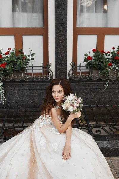 Une belle jeune mariée portant une robe de mariée en dentelle luxueuse posant avec un bouquet de mariée à l'extérieur. Portrait romantique extérieur d'une jolie fille brune avec un maquillage lumineux et une coiffure de mariage — Photo