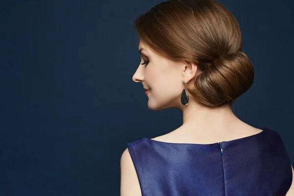 Ung kvinna med ljus makeup och elegant frisyr bär blå klänning poserar på den blå bakgrunden. Ett porträtt av en elegant modell med kväll coiffure över blå bakgrund — Stockfoto