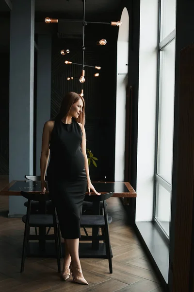Una giovane donna incinta bella in un abito nero sta posando all'interno buio del ristorante e guardando da parte — Foto Stock
