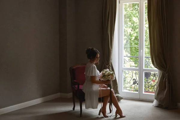 Młoda piękna panna młoda w jedwabnym peignir trzyma bukiet ślubny i siedzi na krześle w pokoju hotelowym — Zdjęcie stockowe