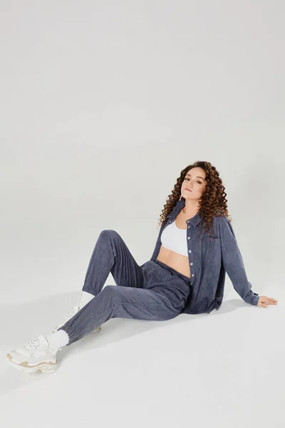 Charming young girl with curly hair dressed in a denim costume and white top posing against a white background. Slim toned model woman in casual clothes is sitting on the floor over white background — 스톡 사진