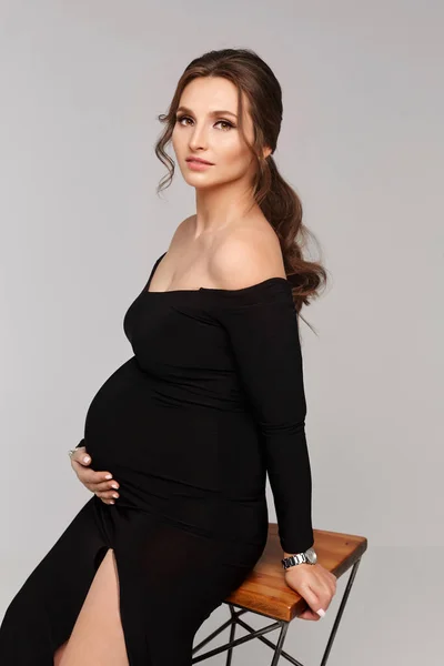 一个穿着时髦晚礼服的怀孕模特儿紧紧抓住肚子，坐在白色的背景上。年轻美丽的孕妇，身穿黑色长裙，灰色背景，孤立无援 — 图库照片