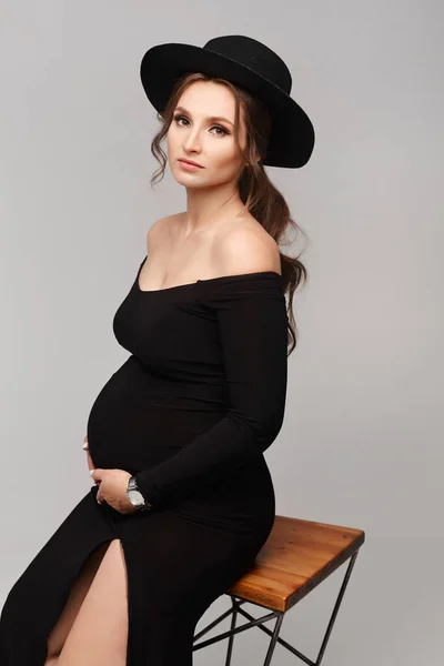 Беременная женщина в длинном платье и большой шляпе, держащая живот на светло-сером фоне — стоковое фото