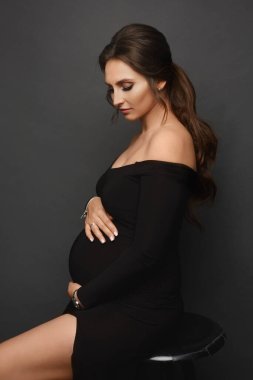 Gri arka planda siyah bir gece elbisesi giyen muhteşem hamile bir kız. Şık giyinmiş hamile bir kadın.
