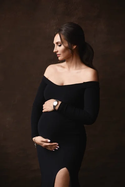 Fashion portrait of a gorgeous pregnant model girl wearing black evening dress posing over brown background — 图库照片