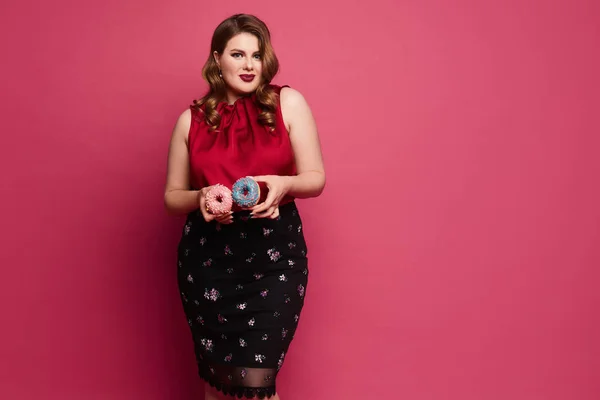 Uma jovem mulher gorda em uma blusa vermelha e saia preta segurando saborosos donuts na mão e olhando para a câmera, isolada no fundo rosa — Fotografia de Stock