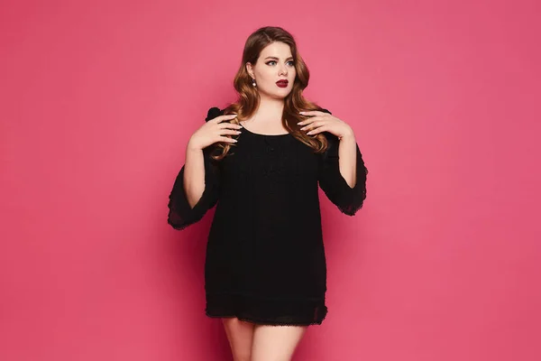 Young shocked plus-size model woman in short dress over pink background. Plump pretty woman with bright makeup in a black dress posing on the pink background, isolated — 图库照片