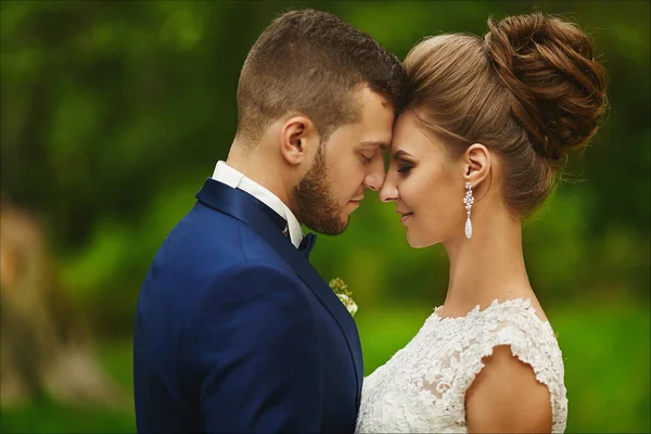 Modish vőlegény megölel egy gyönyörű menyasszonyt az esküvő alatt. Egy szerető pár, egy nő menyasszonyi ruhában és egy jóképű férfi elegáns kék öltönyben, csukott szemmel pózolva. — Stock Fotó