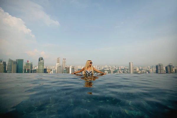 Eine junge hübsche Frau im modischen Badeanzug posiert am Rande des Infinity-Pools auf dem Dach des Gebäudes mit einem Panoramablick auf Singapur — Stockfoto