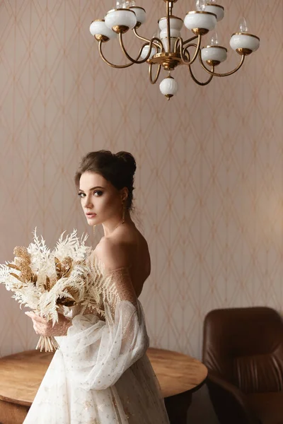 Retrato recortado de uma modelo feminina usando um vestido de noiva vintage com mangas compridas dentro de casa. Elegante jovem noiva em vestido vintage elegante posando com buquê de noiva elegante no interior — Fotografia de Stock