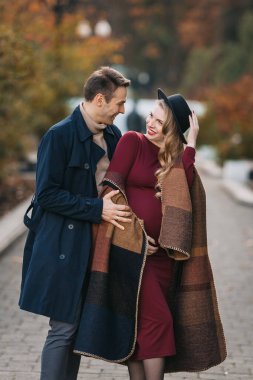 Mutlu genç ebeveynler, yağmurluk giyen şık bir adam ve sonbahar günü bir sonbahar parkında birlikte yürüyen kırmızı elbiseli ve şık şapkalı hamile bir kadın.