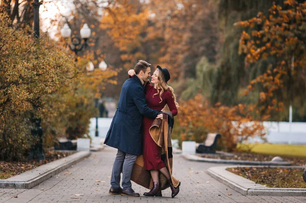 Junges verliebtes Paar, schöner Mann und elegante Frau in rotem Kleid und Hut umarmen sich und haben Spaß in einem Park an einem Herbsttag — Stockfoto