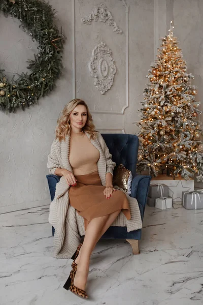 Hermosa mujer rubia joven sexy con ropa elegante sentada en un sillón cerca del árbol de Navidad en el interior decorado para la celebración de Año Nuevo —  Fotos de Stock