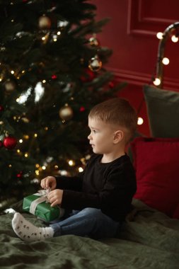 Noel hediyesi paketini süslenmiş bir Noel ağacının yanındaki yatağın üzerine boşaltan sevimli küçük çocuk.