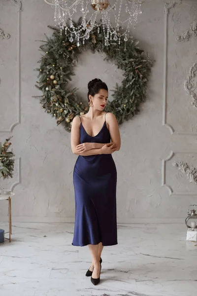 Menina adorável em um elegante vestido azul escuro na frente de decorações de Natal. Modelo feminino adorável em vestido da moda posando no interior decorado para feriados de Ano Novo — Fotografia de Stock