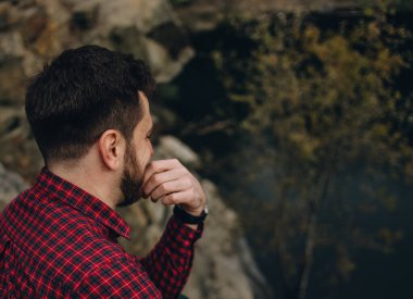 bearded man traveler sits with his back thinking on the shore of a lake with stone