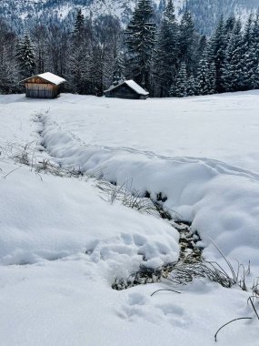Karla kaplı çayır, küçük ahşap kulübeler, çam ormanı ve dramatik bulutlu gökyüzünün altındaki uzak tepeler ile huzurlu bir kış manzarası..