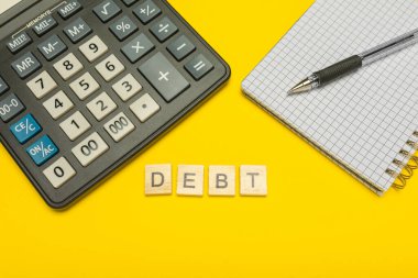 Sözcük borcu sarı arka planda ahşap harflerle ve kalem ve defter ile modern hesap makinesiyle yapılır. Uzayı kopyala Ekonomi, para planlama. İş, finans kavramı. Vergi yükü. Vergiler ve vergiler.